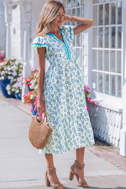 The Meredith Dress: white with blue floral print and turquoise trim, worn outdoors, carrying a woven straw bag.