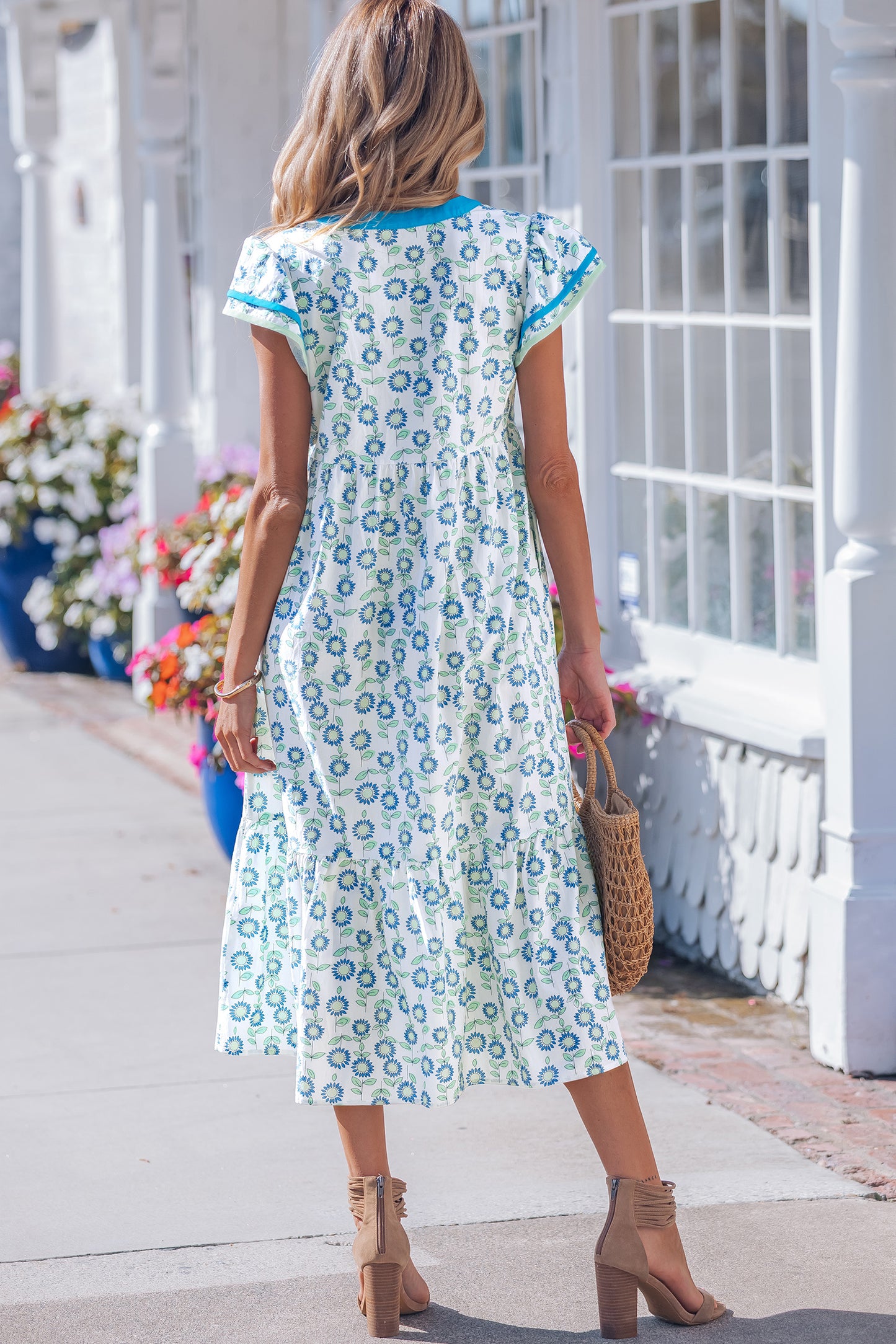 The Meredith Dress in white with blue floral print and blue-trim flutter sleeves, back view with straw bag.